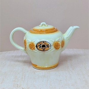 May include: Cream-colored ceramic teapot with a lid and handle. The teapot features orange floral designs and a decorative oval emblem. The lid and base have orange accents. A classic piece for serving tea.