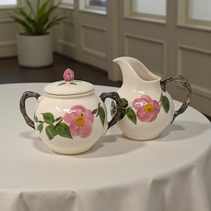 May include: Cream-colored ceramic sugar bowl with lid and creamer set. Each piece is decorated with pink floral designs and green leaves. The handles have a textured, dark brown appearance. The set is on a white tablecloth.