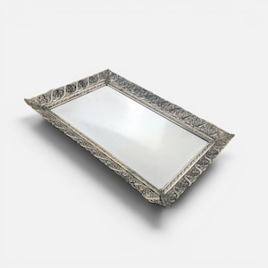 May include: A rectangular silver-toned tray featuring a reflective mirrored base and an ornate, raised border decorated with intricate, repeating floral and vine-like embossed patterns along all four sides of the metallic frame.