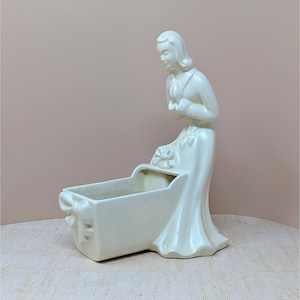May include: Off-white ceramic planter featuring a woman looking down at a rectangular cradle-shaped container. The woman is wearing a long dress and has her hands clasped. The planter has bow details on the cradle.