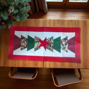 May include: A rectangular Christmas table runner with a red polka dot border. The center features a red star and green and white triangles with holiday-themed fabric. The runner is placed on a wooden table, with a Christmas tree in the background.