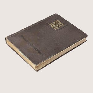 May include: A vintage, brown hardcover book titled "Ruskin's Crown of Wild Olive: The Queen of the Air." The book has a worn appearance with gold-colored page edges. The title is printed in a rectangular box on the cover.