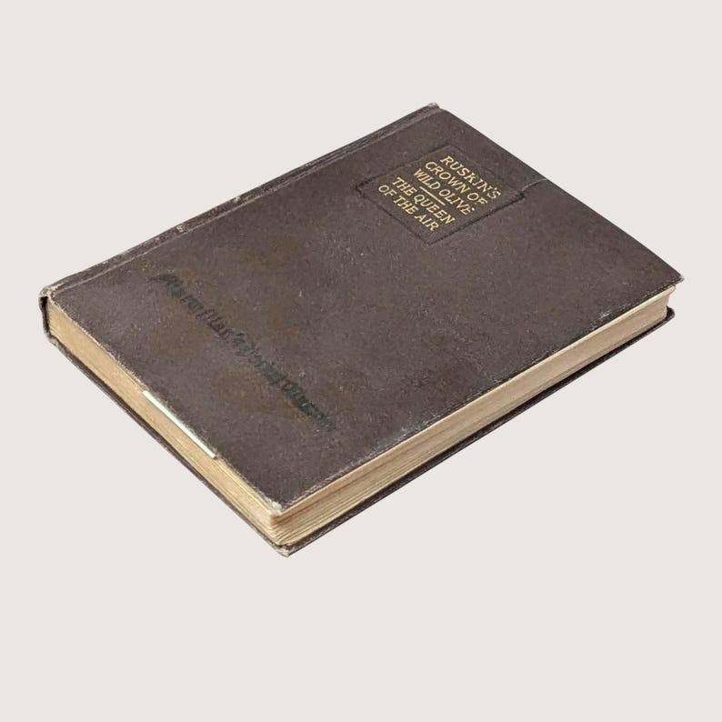 May include: A vintage, brown hardcover book titled "Ruskin's Crown of Wild Olive: The Queen of the Air." The book has a worn appearance with gold-colored page edges. The title is printed in a rectangular box on the cover.