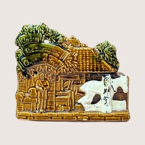 May include: A glazed ceramic decorative plaque featuring a relief scene of a blacksmith shop with a white horse and the text Under the Spreading Chestnut Tree. The piece is finished in shades of brown and green with a glossy, reflective surface.