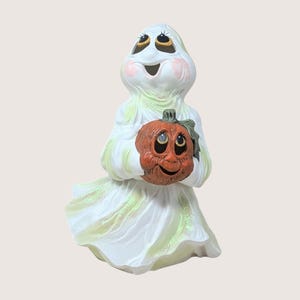 May include: A ceramic Halloween ghost figurine holding a smiling pumpkin. The ghost is white with green accents and rosy cheeks. The pumpkin is orange with black eyes and a smiling mouth. A festive decoration.