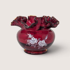 May include: A deep red glass bowl featuring a ruffled, crimped rim and a hand-painted white floral design on the front. The bowl has a rounded body and a circular base, with the white painted roses and leaves contrasting against the dark glass.
