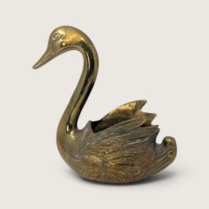 May include: A polished brass swan figurine featuring a long, curved neck and detailed, textured wing feathers. The body of the swan is hollowed out, forming a container with a smooth, reflective surface on the head and neck, and intricate plumage.