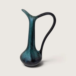 May include: A tall, slender ceramic pitcher with a bulbous base and a long, curved neck. The piece features a deep teal and black glaze with a streaked, vertical finish. It includes a single, smooth black handle extending from the neck to the base.