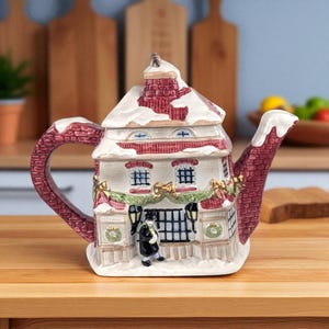 May include: A ceramic teapot in the shape of a house, with a red brick chimney and roof, and snow detailing. The handle and spout are also red brick. A small figure stands at the front, with festive garlands and wreaths. The teapot is designed for tea.