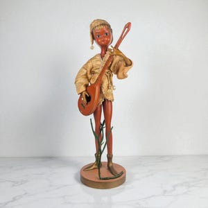 May include: A vintage figurine of a musician in a gold-colored outfit, holding a brown stringed instrument. The figurine has a painted face and is standing on a round base with a decorative branch. The figure is approximately 12 inches tall.