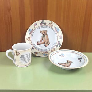 May include: A three-piece children's dish set featuring a teddy bear design. The set includes a plate, a bowl, and a mug. The plate and bowl have a gold trim. The mug has a curved handle and text. The set is cream-colored with a teddy bear illustration.