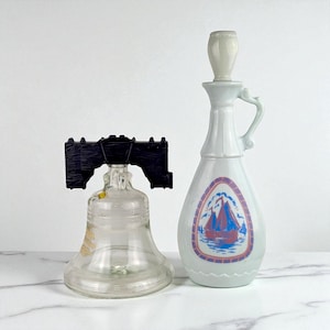 Può includere: Due bottiglie decorative in vetro. Una è a forma di Liberty Bell con un tappo in plastica nera. L'altra è una bottiglia bianca con manico e un disegno di barca a vela in un ovale rosso e blu.