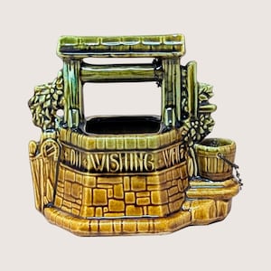 May include: A ceramic wishing well planter featuring a glossy olive green and brown stone-textured finish. The structure includes a roof, a small attached bucket on a chain, and the embossed text "OH WISHING WELL" across the front facade.