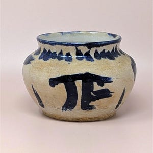 May include: A small, round ceramic pot with a wide opening. The pot is off-white with dark blue brushstrokes and a band of blue floral-like designs around the rim. The pot has a simple, rustic aesthetic.