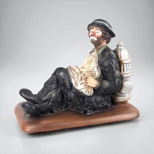 May include: A figurine of a seated clown, dressed in a black suit and hat, holding a newspaper. The clown has a painted red nose and is leaning against a silver fire hydrant. The base is brown.