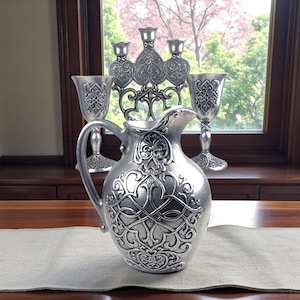 May include: A silver-colored pitcher with an ornate design, accompanied by two goblets and a candelabra. The pitcher has a handle and a detailed pattern. The goblets and candelabra share a similar design. The items are displayed on a wooden surface with a neutral-colored runner.