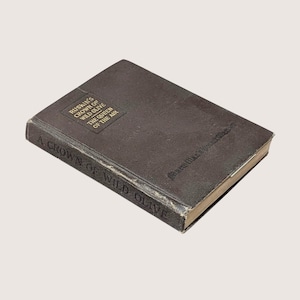 May include: A vintage book titled "A Crown of Wild Olive" by Ruskin. The dark brown cover features gold lettering and a rectangular emblem with text. The book's edges show signs of wear, suggesting its age and use.