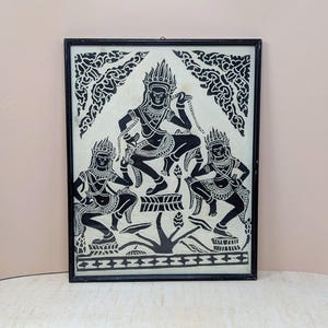 May include: Black and white framed artwork depicting three figures in traditional attire. The central figure is seated, flanked by two others, all with ornate headdresses. The artwork has a detailed, symmetrical design, with a cream-colored background.