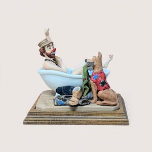 May include: A hand-painted figurine of a clown with a red nose and brown hat sitting in a light blue bathtub. A tan dog with a red and blue patched vest sits beside the tub, holding a green cloth. Clothing items are piled on the wooden base.