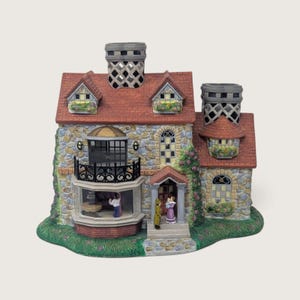 May include: A ceramic miniature house figurine featuring a stone-textured facade, a red shingled roof, and two lattice-patterned chimneys. The structure includes a balcony, flower boxes, and two small figures standing near the front entrance.