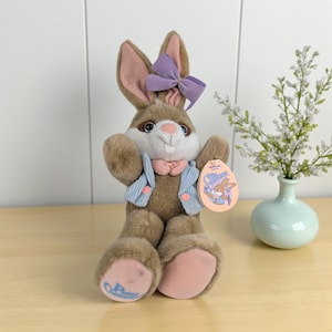 May include: A plush brown bunny with a lavender bow, pink ears, and a blue vest. The bunny has a pink nose and paws. A tag reads "Peter Cottontail". The bunny is a stuffed animal.