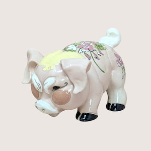 Puede incluir: Figura de cerdo de cerámica rosa claro, adornada con motivos florales rosas y verdes. El cerdo tiene pezuñas negras, una mancha amarilla en la cabeza y detalles blancos alrededor del hocico y las orejas. Artículo decorativo.