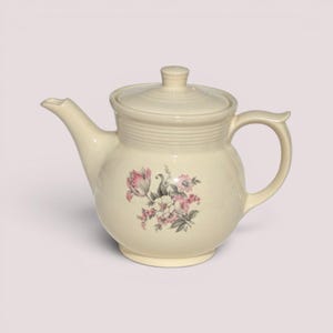 May include: A cream-colored ceramic teapot featuring a rounded body, a curved spout, and a matching lid. The center of the teapot is decorated with a floral illustration in shades of pink, white, and grey, depicting various blossoms and leaves.
