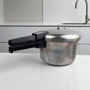 Könnte beinhalten: Ein Schnellkochtopf aus Edelstahl mit schwarzem Griff. Der Topf hat einen silbernen Deckel und ein abgenutztes Aussehen. Der Kocher steht auf einer weißen Oberfläche, mit einem Küchenbecken im Hintergrund.