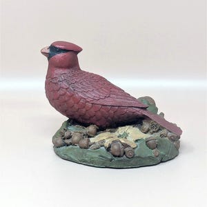 Puede incluir: Figurilla detallada de un pájaro cardenal rojo con cara y cresta negras. El pájaro está posado sobre una base con bellotas y hojas, pintadas en tonos verdes y marrones. La figurilla es un objeto decorativo.