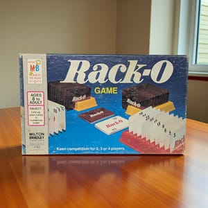May include: A vintage Rack-O game box with a blue background and the game's title in white. The box features illustrations of game components, including cards and racks. The game is for ages 8 to adult and for 2, 3 or 4 players.