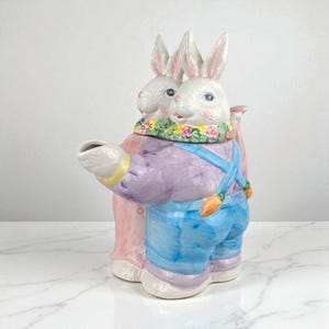 May include: A ceramic teapot in the shape of two white bunnies. One bunny is wearing a purple shirt and blue overalls with carrot accents. The other bunny is wearing a pink coat. A floral garland adorns their necks.