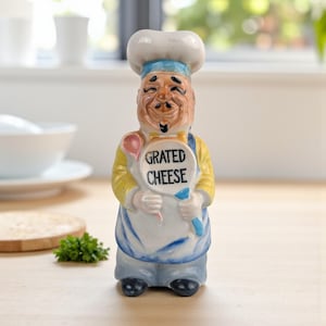 May include: A ceramic chef figurine with a smiling face, wearing a white hat, blue and white apron, and yellow shirt. The figurine holds a sign that reads "GRATED CHEESE". A pink spoon and blue utensil are also visible.