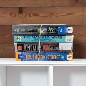 Può includere: Una pila di quattro libri di fantascienza legati con un elastico. I titoli visibili sono "Polaris", "The Man-Kzin Wars", "Enemies" e "Starfar". I libri sono su una mensola bianca contro uno sfondo di legno.