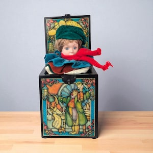 May include: A black, rectangular music box with a hinged lid, featuring a doll with a green hat and red scarf. The box is decorated with a colorful illustration of people and the words "Merry Christmas".