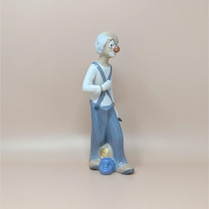 May include: A porcelain clown figurine. The clown is wearing a white shirt, blue overalls, and a red nose. The figurine has a sad expression and is holding a small object. A blue clown head is at the base.