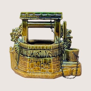May include: A glazed ceramic wishing well planter in shades of olive green and brown. The structure features a brick-patterned base with the embossed text WISHING WELL, a roofed top, sculpted foliage on the sides, and a small bucket on a metal chain.