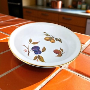 May include: White ceramic serving bowl with a gold rim and fruit illustrations. The bowl features images of plums, pears, and other fruit. The bowl is sitting on a red-orange tiled surface.