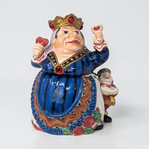 May include: A ceramic figurine of the Queen of Hearts from Alice in Wonderland. The queen wears a gold crown with red jewels, a blue and white dress, and holds heart-shaped objects. A smaller figure stands beside her.