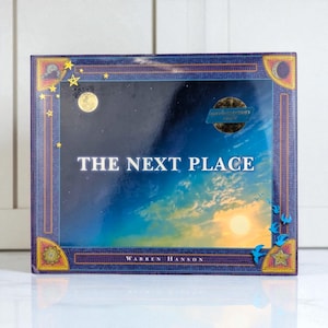 May include: A hardcover book titled "THE NEXT PLACE" by Warren Hanson. The cover features a night sky with a full moon and stars, transitioning to a daytime sky with a setting sun. The border is dark blue with decorative elements.
