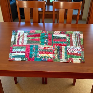 Può includere: Runner da tavolo rettangolare in patchwork a tema natalizio. Il runner presenta una varietà di tessuti rossi, verdi e bianchi, disposti in un motivo geometrico. Il runner è posto su un tavolo di legno marrone.