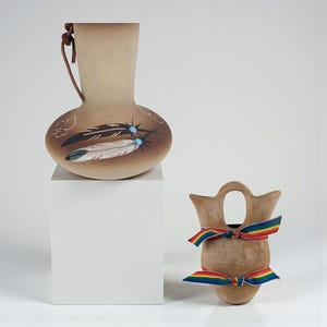 Puede incluir: Dos jarrones de cerámica. El primero es un jarrón marrón en forma de calabaza con un asa de cuero y diseños de plumas y nubes. El segundo es un jarrón más pequeño con una cinta arcoíris atada. Ambos jarrones están sobre un fondo blanco.