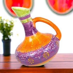 May include: A colourful ceramic jug with a curved spout and handle. The jug is hand-painted with orange, purple, green, and yellow hues. It features decorative swirls and dots, creating a unique and artistic appearance. A decorative item.
