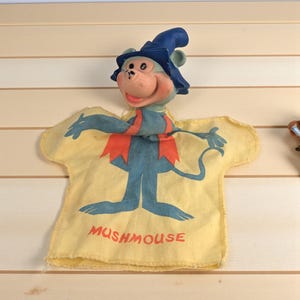 May include: A vintage Mushmouse puppet with a blue monkey head wearing a blue hat. The puppet's body is a yellow cloth with a blue monkey design and the word "Mushmouse" in red.