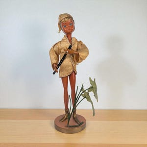 May include: A vintage doll with a tan complexion, wearing a gold-colored outfit and holding a black flute. The doll has blue eyes and is standing on a round base with a plant. The doll is approximately 12 inches tall.