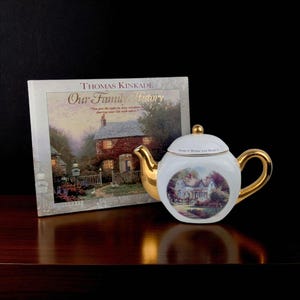 May include: A white teapot with gold accents, featuring a scenic painting and the text "Home Is Where The Heart Is." Beside it is a book titled "Our Family History" by Thomas Kinkade.