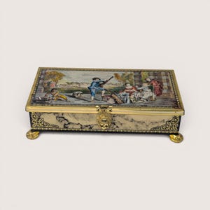 May include: A rectangular box with a hinged lid featuring a colorful pastoral scene of figures in period clothing. The sides have a cream and black marbled finish with gold-toned metal filigree corners, a central latch, and four small feet.