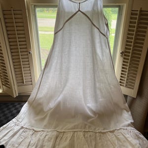 Puede incluir: Un vestido lencero blanco sin mangas con tirantes finos, una costura en forma de V en el corpiño y un volante ancho fruncido en el dobladillo. La prenda está confeccionada en un tejido ligero e incluye un delicado borde de encaje.