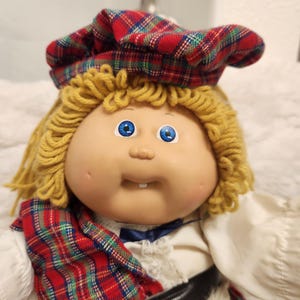 May include: A doll with yarn-like blonde hair, bright blue eyes, and a single visible tooth. The doll wears a red, green, and blue plaid tam-o'-shanter hat, a white shirt with a small blue bow, and a black belt over a plaid skirt with white trim.