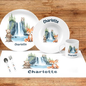 May include: A children's dinnerware set featuring a whimsical forest scene with a waterfall, fox, bear, and owl. The set includes a plate, bowl, mug, and placemat, all with the name "Charlotte" printed on them. A spoon and fork are also included.