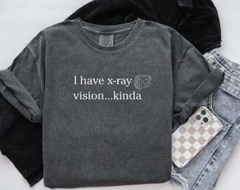 May include: Dark gray long-sleeve shirt with the text "I have x-ray vision...kinda" in white. The shirt has a crew neck and a relaxed fit. A phone with a checkered case rests on a pair of blue jeans.